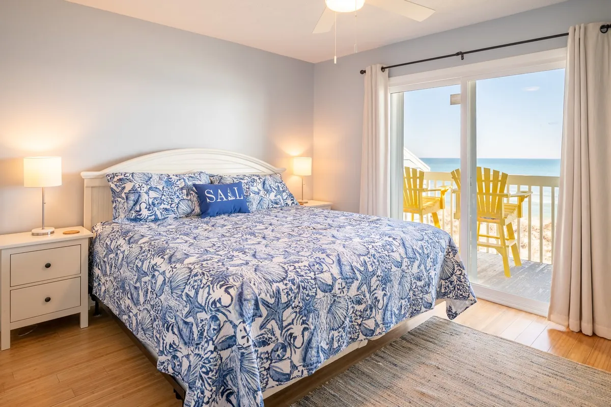 The image showcases a bright, inviting bedroom with crisp blue and white bedding, a large sliding glass door opening to a balcony with cheerful yellow chairs, and sweeping ocean views just beyond. Natural light fills the space, and the ocean horizon is visible from the comfort of the bed, suggesting a tranquil and rejuvenating retreat.