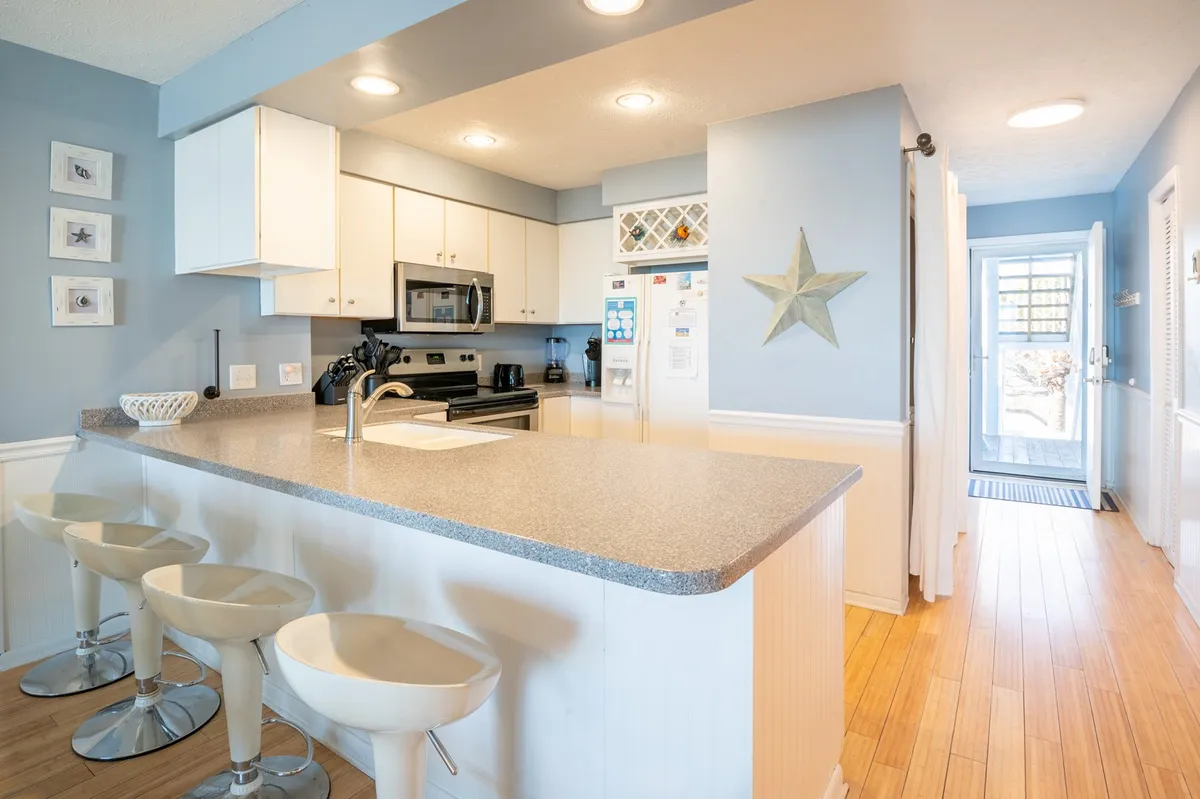 A bright kitchen featuring a prominent island with four bar stools, white cabinetry, and a star decoration. The island serves as a casual dining or prep space, echoing the section’s mention of a welcoming island with seating.