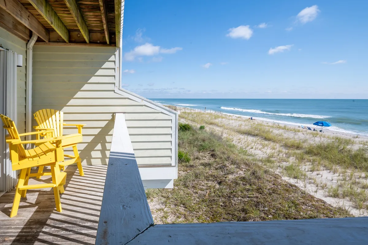 Yellow Adirondack chairs sit atop a wooden deck overlooking sand dunes and the ocean, creating a peaceful and inviting outdoor scene. The area suggests a space for sunbathing, reading, or simply soaking in the natural beauty of the seaside environment.