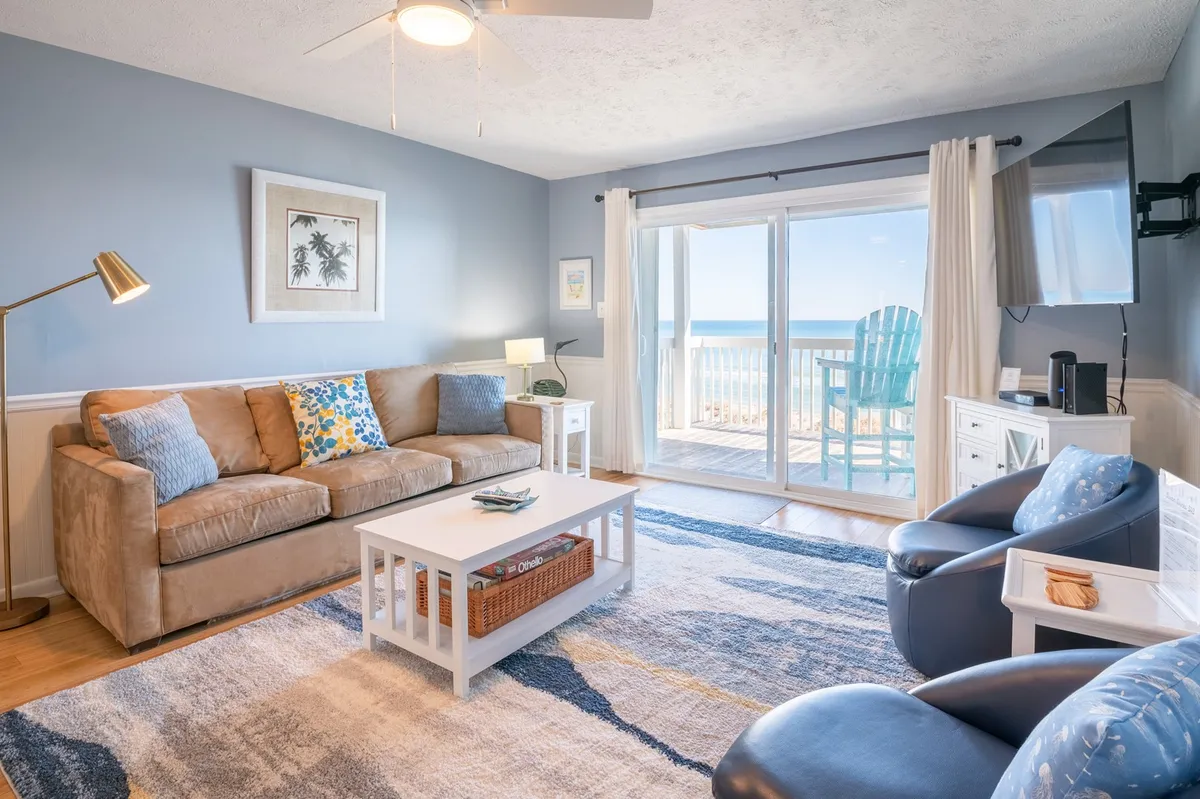 A bright, open-concept living room decorated in soothing coastal pastels. The space features a plush sofa (which likely serves as a Queen Sofa Sleeper), a coffee table, and accent chairs facing a large TV. Through sliding glass doors, you can see blue chairs on a private balcony with sweeping ocean views, letting in abundant natural light.