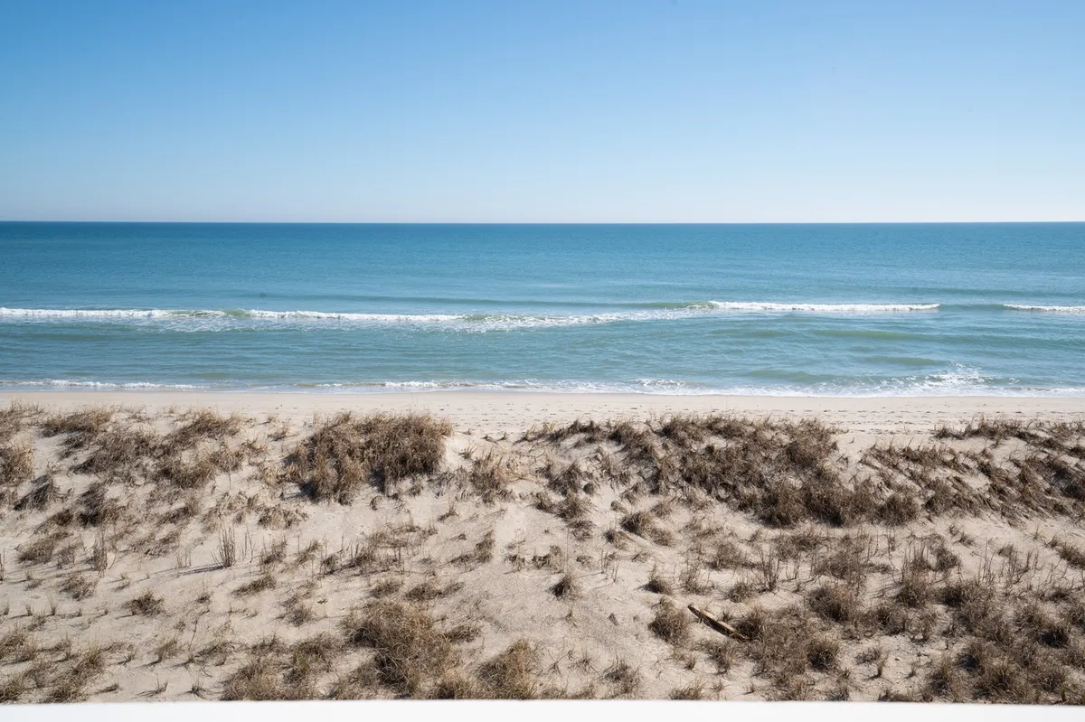 A sweeping view of the sandy beach with gentle ocean waves rolling onto the shore, bordered by dune grasses. The open expanse suggests space for building sandcastles, beachcombing, and enjoying family time by the water.