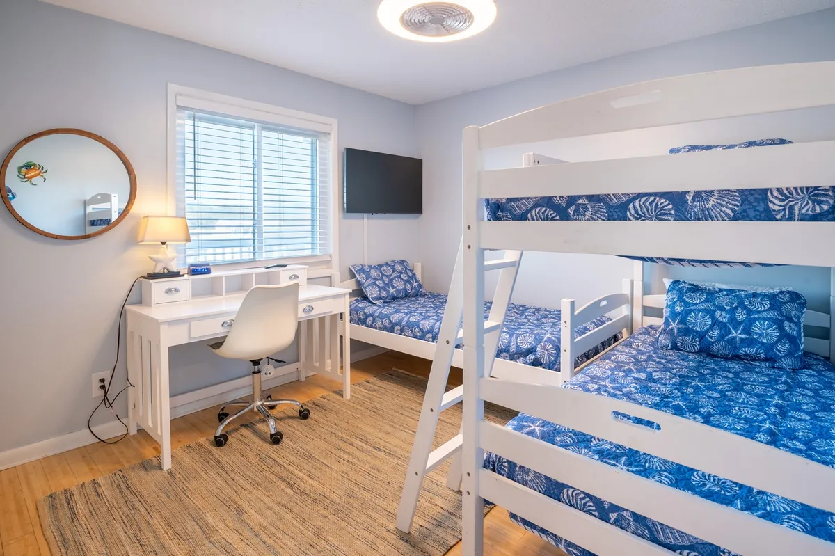 A playful bunk room with twin over twin bunk beds, a standalone twin bed, a desk area, mirror, and television. The space is bright and designed for both sleep and activities, ideal for kids or extra guests.