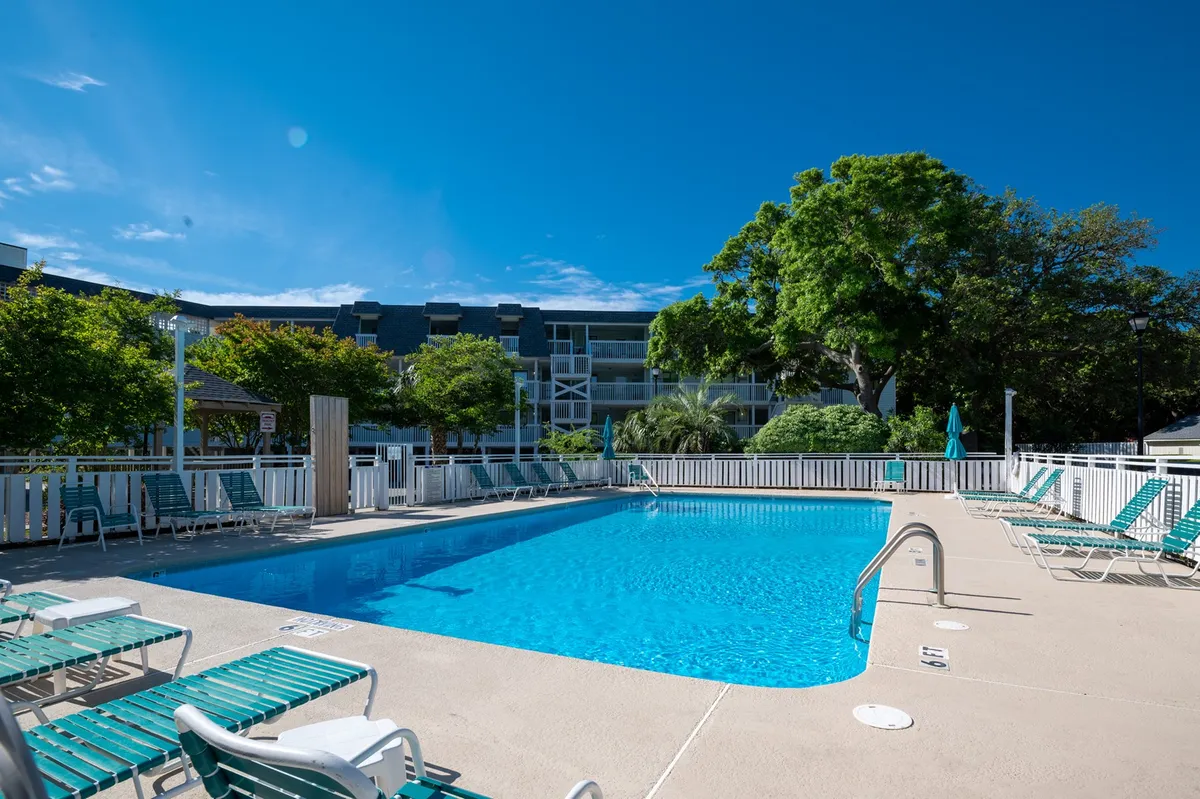This exterior image features a large swimming pool surrounded by lounge chairs and lush greenery under a clear blue sky. The relaxed and cheerful mood is emphasized by people sunbathing, swimming, and socializing, capturing the essence of resort life and communal outdoor enjoyment.