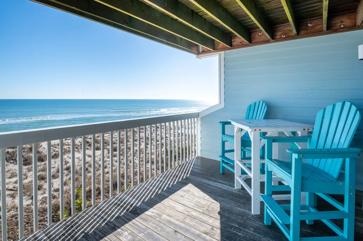 A private balcony with turquoise chairs and a table overlooks the sweeping ocean, inviting guests to savor the tranquil seaside views. The vantage point perfectly captures the early morning light, creating a serene spot for sipping coffee or enjoying a romantic meal while listening to the waves.