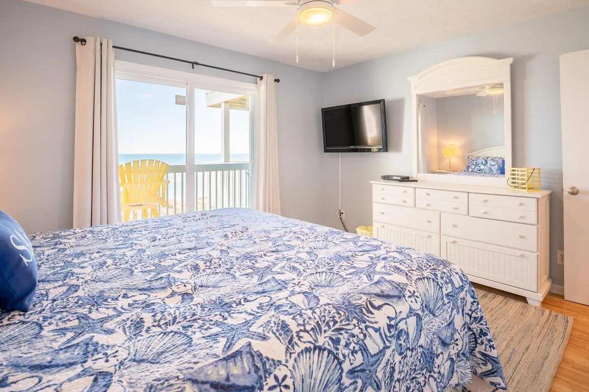 A serene bedroom with a blue and white patterned king bed, large window offering an ocean view, and bright yellow chairs on a private balcony. A white dresser with a mirror sits opposite the bed, completing the restful, beachy ambiance.
