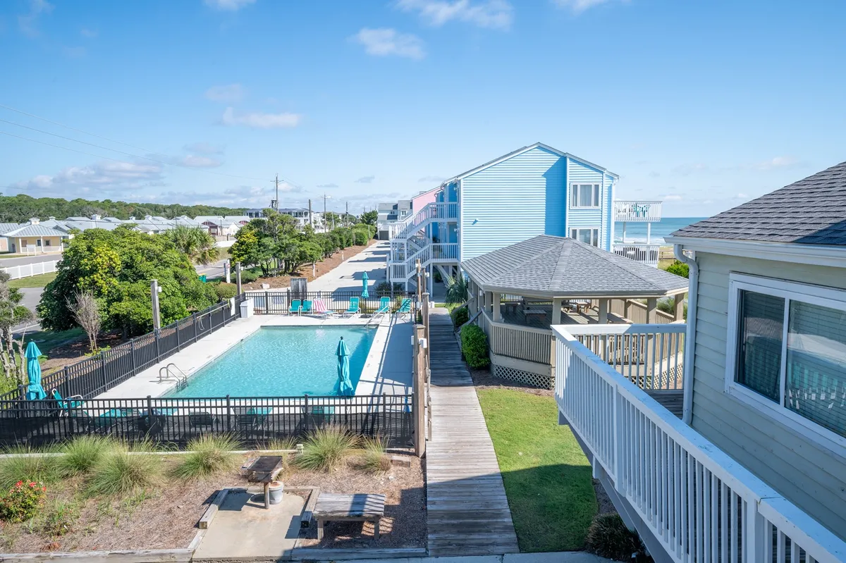 An exterior shot of the Ocean Dunes Resort, focusing on a sparkling swimming pool surrounded by blue resort buildings and lush green landscaping. The area is set up for swimming, sunbathing, and social gatherings, with a relaxed mood enhanced by sunset lighting.