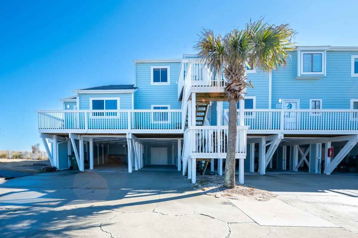 This image features a cheerful exterior scene with a palm tree, a staircase leading to a balcony, and blue siding. The focal points are the balcony and outdoor seating spaces, suggesting a vibrant area perfect for gatherings, barbecues, and relaxing with friends. The setting is bathed in natural light, evoking a sense of warmth and camaraderie.
